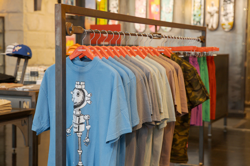 A clothing rack of folded t-shirts inside Unheardof Brand, with skateboard decks displayed on the wall behind.