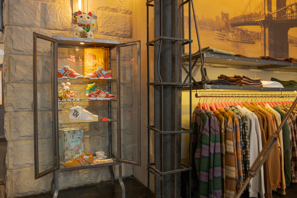 A glass display case of sneakers and collectible toys next to a framed photo of the Cincinnati riverfront and a rack of flannel shirts.