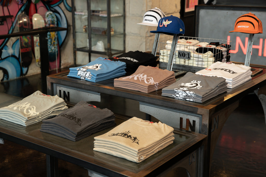 Folded Unheardof-branded t-shirts and caps on display tables, with skateboard decks and mural artwork behind.